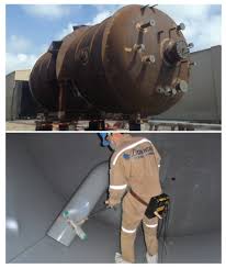 A steel process tank above, and below it an operator in protective clothing spraying its lining.